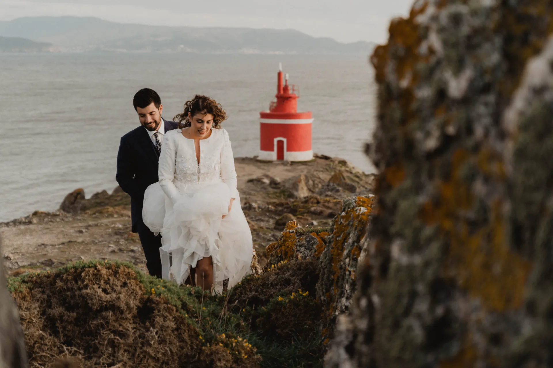 Novios sentados en las rocas junto a un faro rojo